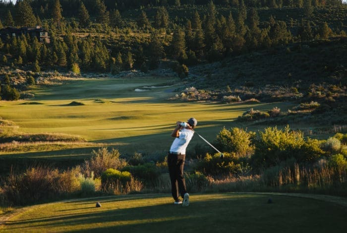 a Golfer in a White Shirt and Dark Pants Swings on a Tee Box Facing a Sunlit Fairway at a Bend Oregon Golf Course the Lush Landscape is Surrounded by Green Shrubs Trees and Rolling Hills with a Building Nestled Among the Trees