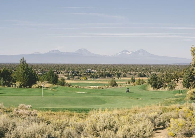 the Scenic Golf Course at Brasada Ranch with Lush Green Fairways and a Golf Cart Sits in the Center Sagebrush and Trees Surround the Course Near Rolling Forests and Snow capped Mountainsperfect for Those Seeking Where to Stay in Bend Oregon