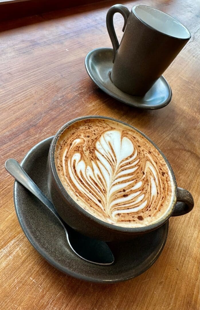 a Cup of Latte with Intricate Leaf shaped Art Sits on a Brown Saucer with a Spoon on a Wooden Table in the Background an Empty Matching Cup Hints at Cozy Moments Found in Restaurants in Frederick Md All Warmly Lit by Natural Light