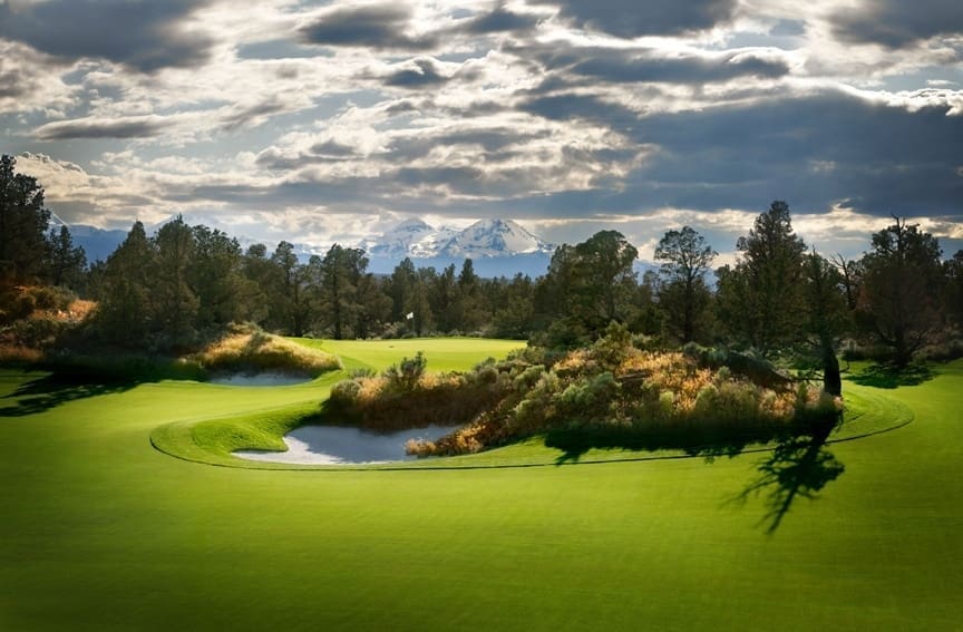 a Lush Green Bend Oregon Golf Course with a Sand Bunker is Surrounded by Trees and Shrubs Under a Dramatic Cloudy Sky Snow Capped Mountains Rise in the Background with Sunlight Filtering Through the Clouds and Casting Shadows Across the Landscape
