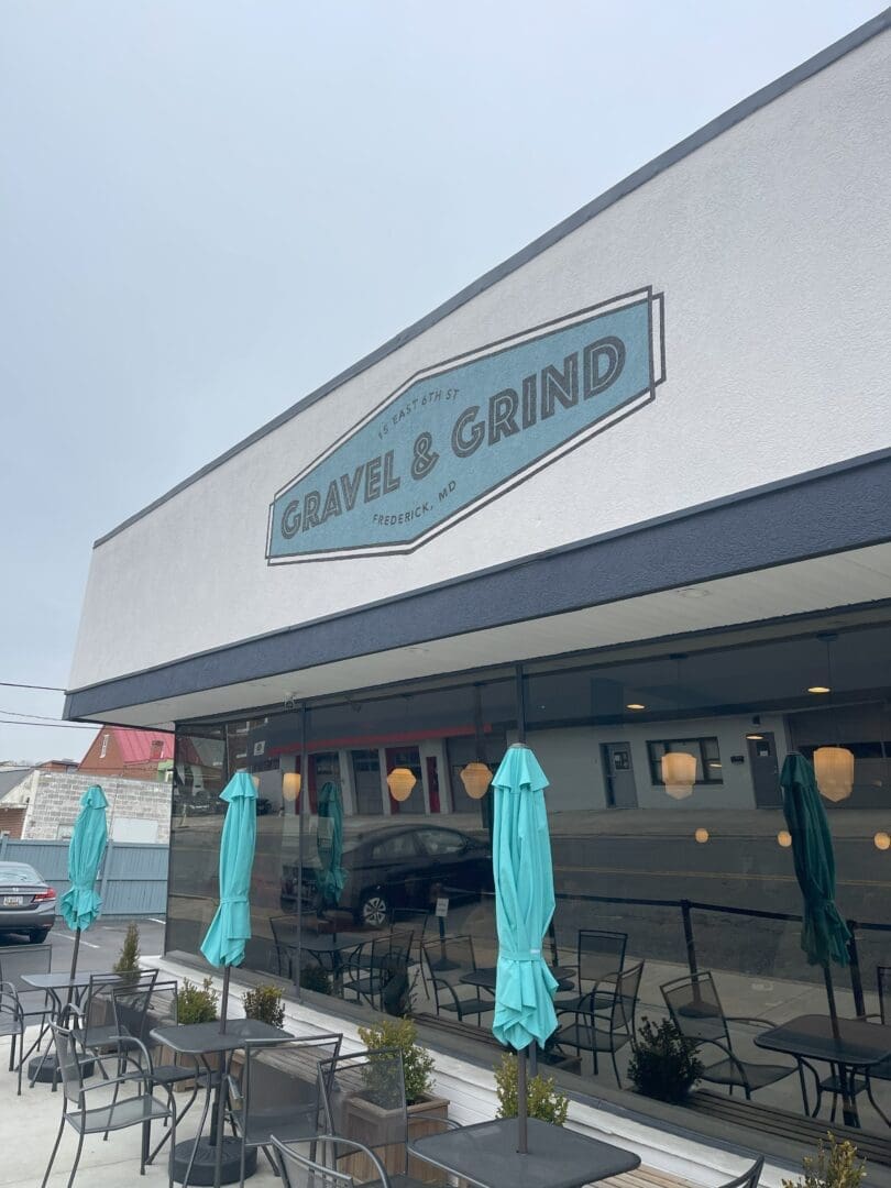 a Modern Cafe Exterior Part of the Vibrant Restaurants in Frederick Md Features a Sign Reading Gravel  Grind Above Large Windows Closed Teal Umbrellas Shade Outdoor Tables with Reflections of Nearby Buildings Visible on a Cloudy Day