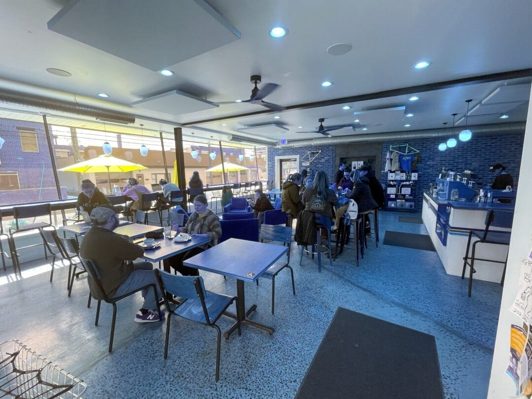 Gravel and grind interior a Bright Modern Coffee Shop in Frederick Md with Large Windows Letting in Sunlight and Yellow Patio Umbrellas Outside People Wearing Masks Sit at Blue Tables and Chairs While a Person Stands Behind a Blue Accent Wall at the Counter