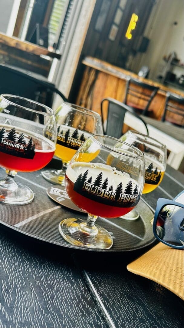 a Close Up of a Tray with Four Stemmed Glasses of Assorted Beer at One of the Best Breweries in Frederick Md Each Bearing the Elder Pine Logo the Rustic Modern Brewery Features Wood and Metal Décor with Drinks Ranging from Red to Amber and Yellow