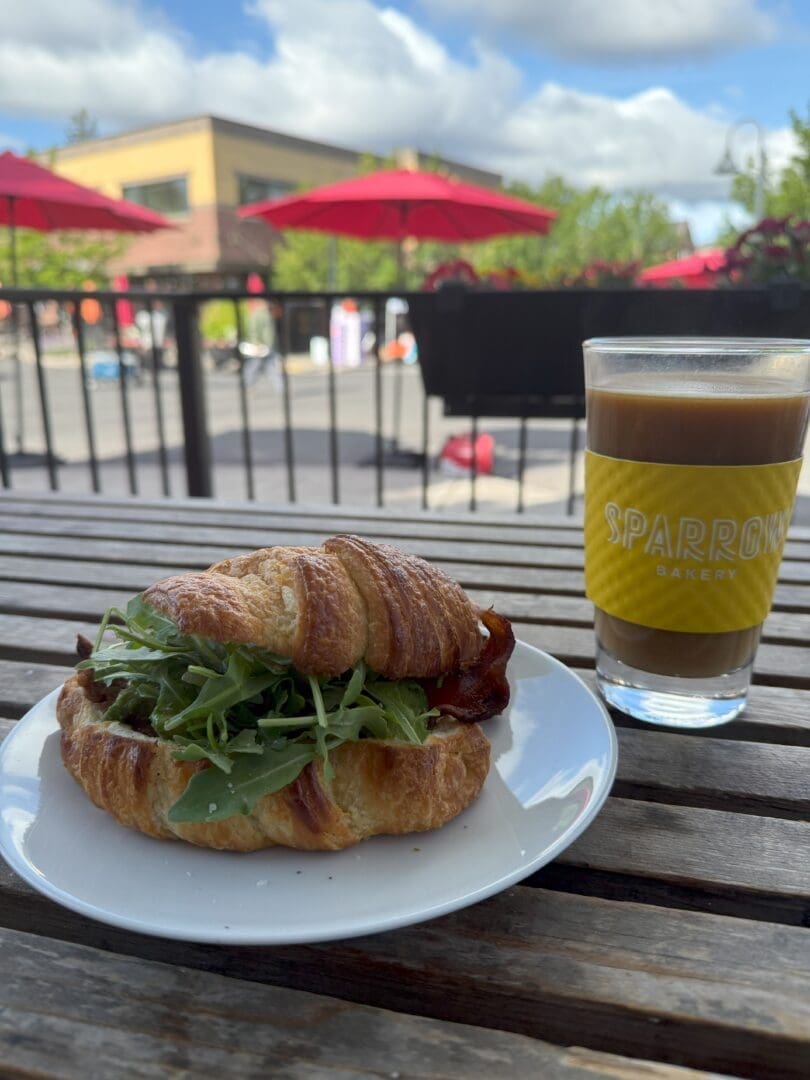 a Croissant Sandwich Filled with Arugula Sits on a White Plate Next to a Glass of Iced Coffee Labeled Sparrow Bakery Atop a Wooden Table Outside Red Umbrellas Greenery and Buildings Are Visible in the Sunny Background