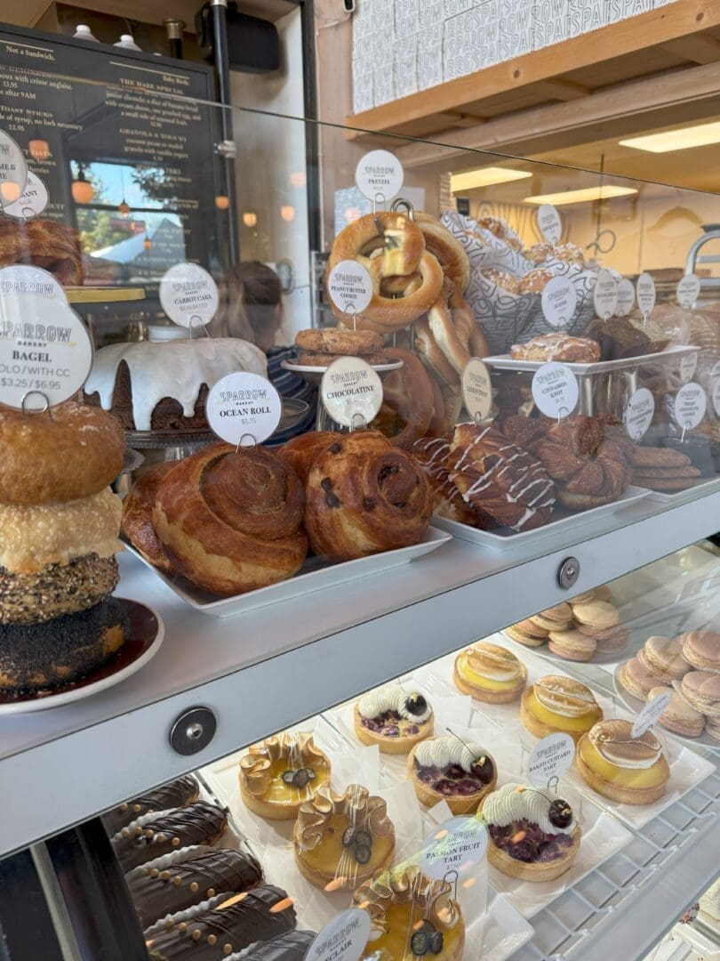 a Bakery Display Case Filled with Pastries Including Stacked Donuts Cinnamon Rolls Braided Breads and Croissants on the Top Shelf with Tarts and Cakes Topped with Fruit and Whipped Cream on the Lower Shelf Labels Mark Each Item
