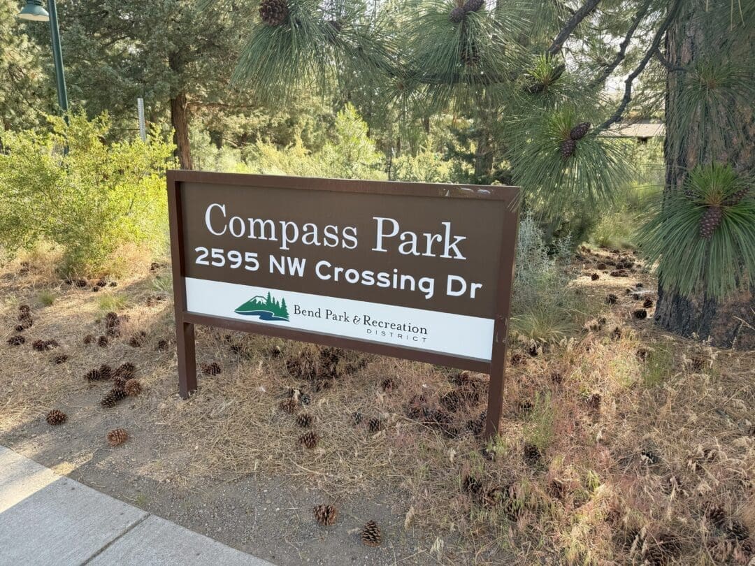 a Brown and White Sign Reads Compass Park 2595 Nw Crossing Dr with a Green Park Logo Below Standing Amid Pine Trees Dry Grass and Scattered Pinecones the Sidewalk is Visible on the Left