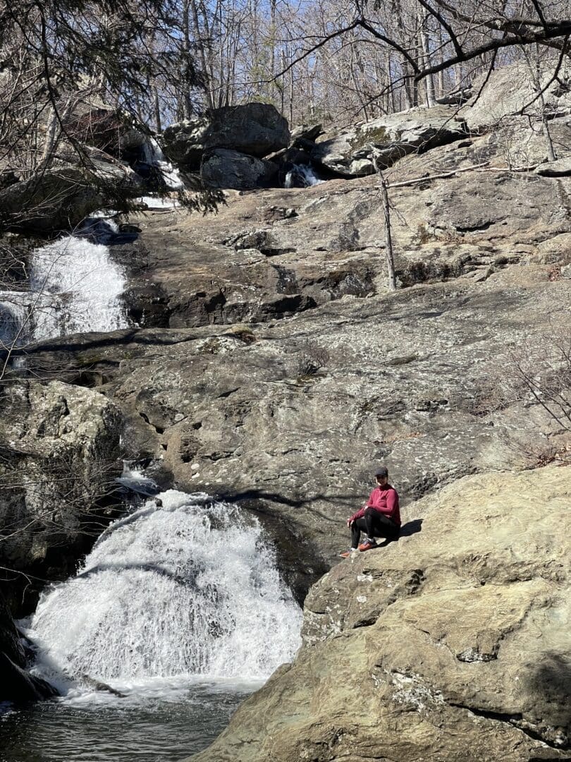 a Person in a Red Jacket Sits on a Large Rock at Cunningham Falls State Park Facing a Small Waterfall Cascading over Rugged Terrain Leafless Trees and Scattered Boulders Surround the Scene Suggesting Early Spring or Late Fall