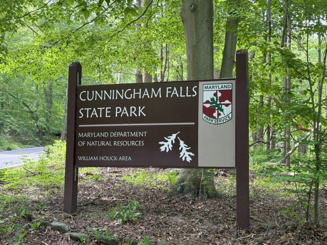 a Wooden Sign for Cunningham Falls State Park Stands in a Forested Area Near Mt Airy Md the Sign Features the Maryland Park Service Logo White Oak Leaves and Text for the Maryland Department of Natural Resources William Houck Area Lush Green Trees Surround It