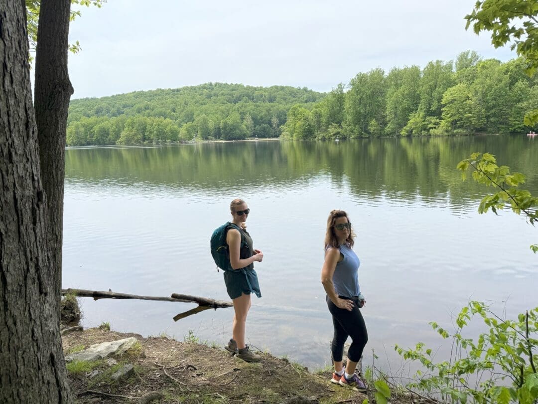 Two Women Stand by a Calm Lake at Cunningham Falls State Park Surrounded by Lush Green Trees and Hills One Wears a Backpack and Shorts the Other Leggings and Sunglasses Both Face the Camera with Reflective Water and Forested Shoreline Behind Them