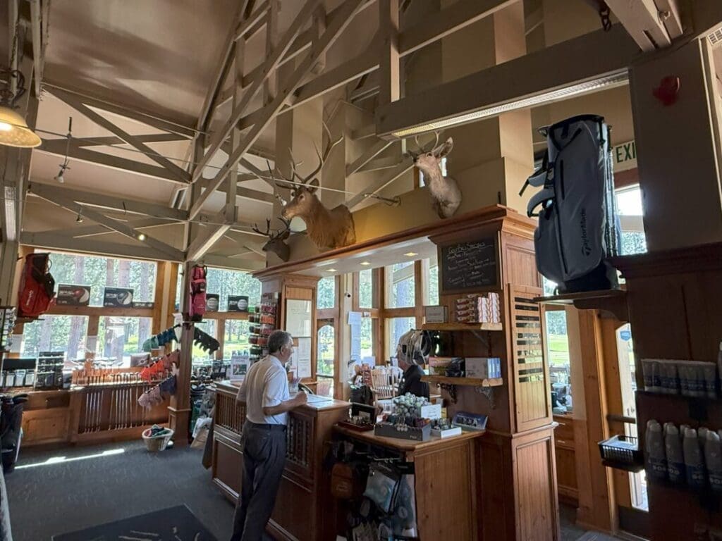 a Man Stands at a Wooden Counter in a Golf Pro Shop with Large Windows Behind the Counter a Woman Assists Him Golf Gear Apparel and Displays Fill the Shop Two Deer Mounts Are on the Beam Above and Sunlight Streams Through the Tall Windows