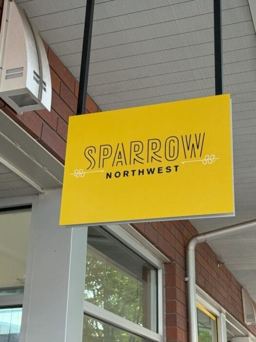 A yellow hanging sign with “Sparrow Northwest” in bold black letters and simple decorative lines, mounted under a covered walkway with a red brick wall and windows in the background.