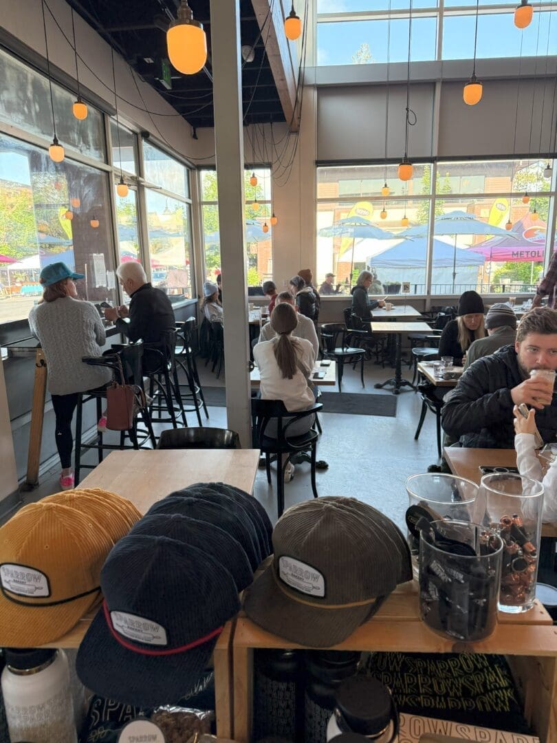 a Bright Modern Café with Large Windows and Hanging Lights Where People Sit at Tables and Bar Seating Chatting or Working in the Foreground Hats Cups and Coffee Beans Are Displayed on a Wooden Counter Outdoor Seating and Umbrellas Are Visible Outside
