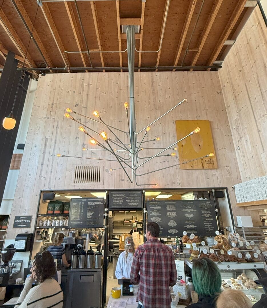 a Modern Cafe Interior with Light Wood Walls an Industrial Chandelier with Exposed Bulbs and a High Unfinished Ceiling Customers Stand at the Counter Baked Goods and Menus Are Visible a Yellow Abstract Painting Hangs on the Back Wall