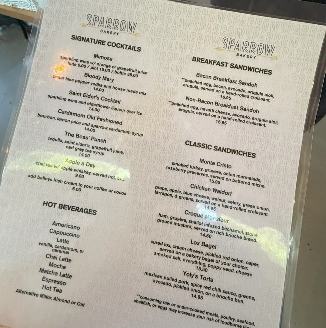 a Laminated Café Menu on a Table Lists Signature Cocktails Hot Beverages Breakfast Sandwiches and Classic Sandwiches Items Include Mimosas Bloody Mary Café Drinks Bacon and Egg Sandwiches Monte Cristo Chicken Waldorf Croque Monsieur and Lox Bagel