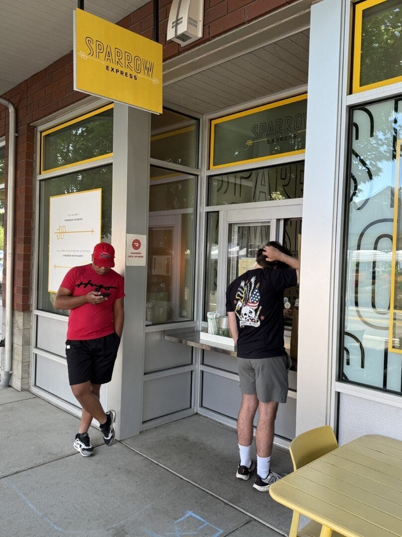 Two People Wait Outside Sparrow Bakery One in a Red Hat and Shorts Looks at His Phone and Leans Against the Wall the Other in a Black Shirt with a Colorful Back Design Stands by the Order Window Yellow Chairs and Tables Are Nearbyperfect for an Ocean Roll Break