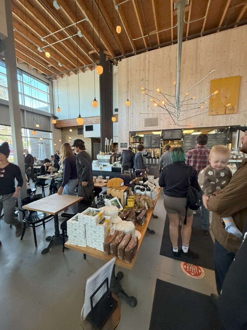 a Busy Modern Café with High Ceilings Hanging Lights and Large Windows People Wait in Line at the Counter and Sit at Tables Breads and Packaged Goods Are Displayed on a Central Table a Man Holds a Baby in the Foreground Bright Lively Atmosphere