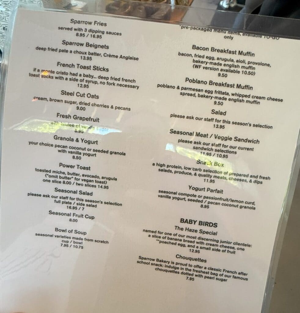 a Printed Cafe Menu on a White Background Lists Breakfast and Brunch Items Such As Sparrow Fries French Toast Sticks Steel Cut Oats Granola Toast Options Muffins Sandwiches Salad Yogurt Parfait Soups and Kids Meals Reflections Are Visible on the Menu