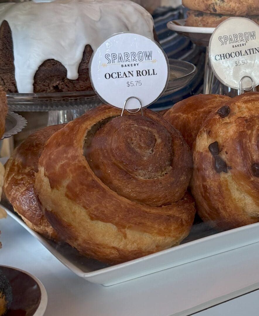 a Close up of a Golden brown Pastry Labeled ocean Roll 5 from Sparrow Bakery Displayed on a White Tray the Pastry is Swirled and Flaky Sitting Beside Other Baked Goods Including a Chocolate Pastry and a Cake with White Icing
