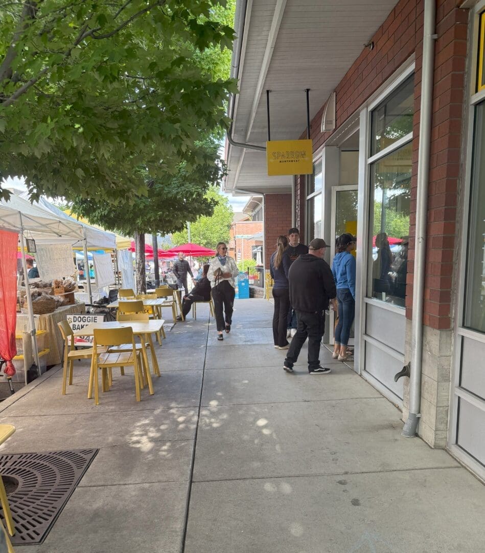 People Stand in Line Outside a Storefront with a Yellow Sign Yellow Chairs and Tables Are on the Sidewalk Under a Tree to the Left Vendors with White Tents and Red Umbrellas Are Set Up at a Market Its a Sunny Day with Green Trees Along the Walkway