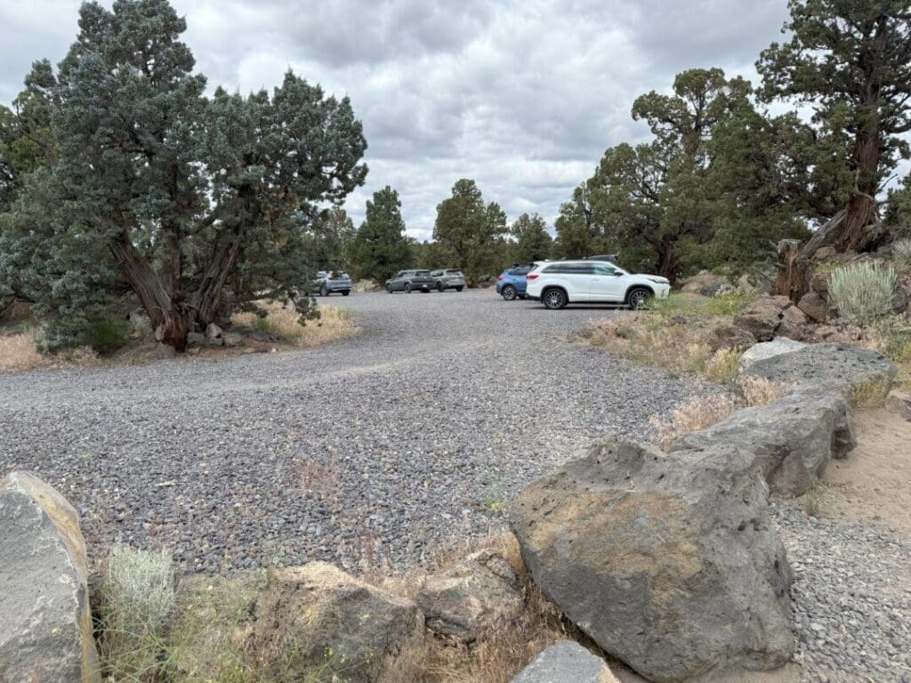 a Gravel Parking Lot with Several Cars Parked Among Scattered Juniper Trees Large Rocks Line the Front Edge and Sparse Grass and Brush Grow Around the Area the Sky is Cloudy Giving the Scene a Muted Overcast Look