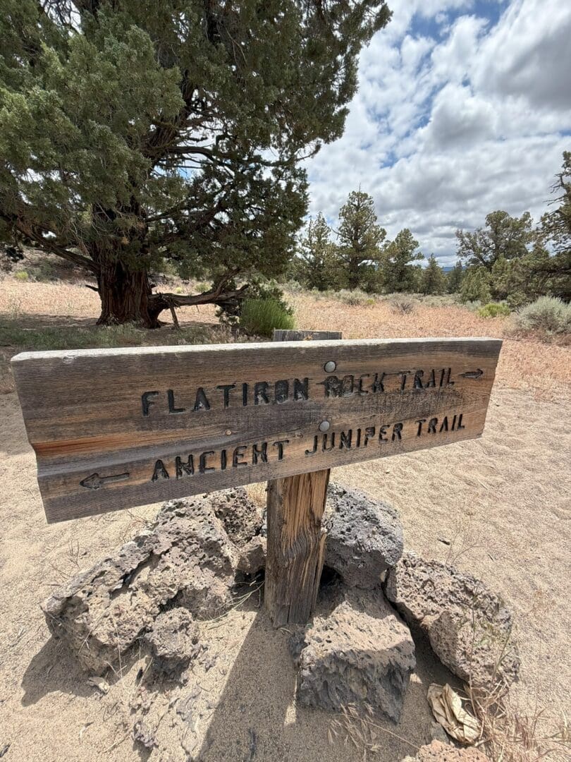 a Weathered Wooden Trail Sign Stands in Sandy Terrain Surrounded by Rocks It Points Left to Flatiron Rock Trail and Right to Ancient Juniper Trail Juniper Trees and Dry Grass Are Visible Under a Partly Cloudy Sky in the Background