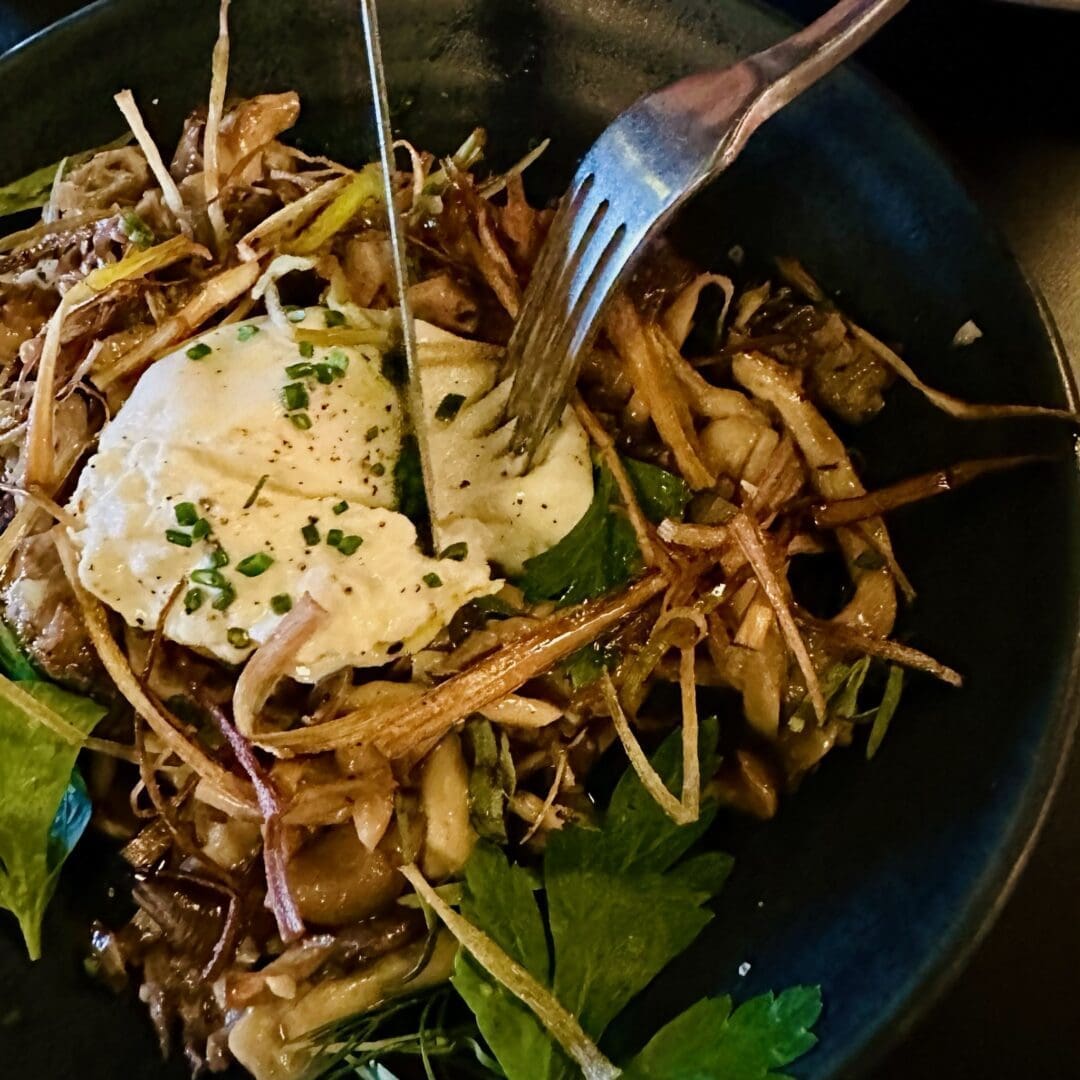 a Dark Bowl Filled with Sautéed Mushrooms Crispy Fried Thin Root Vegetables Fresh Herbs and Topped with a Runny Poached Egg Which is Being Pierced by a Fork the Dish Has an Earthy Rustic Presentation with Vibrant Greens