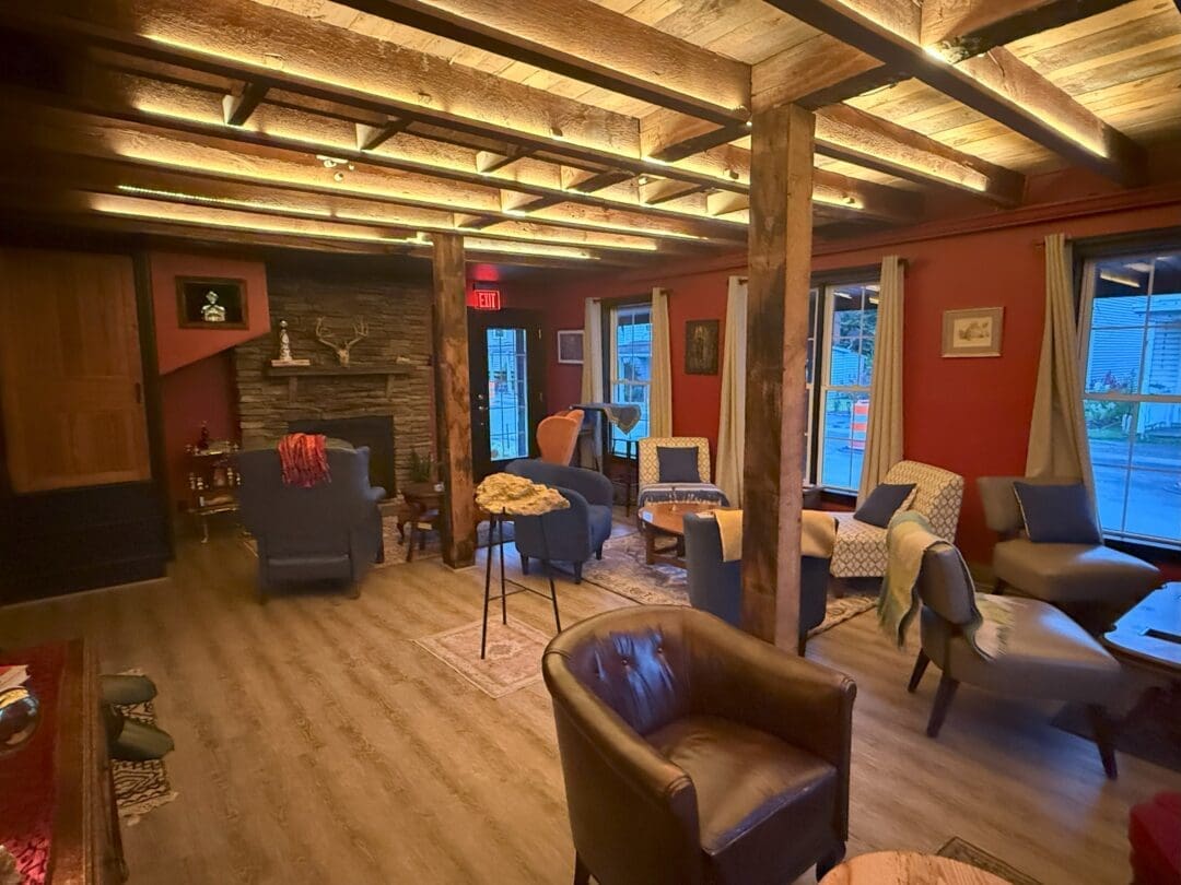 a Cozy Lounge at Market Table Bistro Features Wooden Beams Red Walls and Floor Lamps Armchairs and Small Tables Sit on Rugs a Stone Fireplace and Bookshelves Add Warmth While Large Windows and Draped Blankets Create a Welcoming Relaxed Atmosphere
