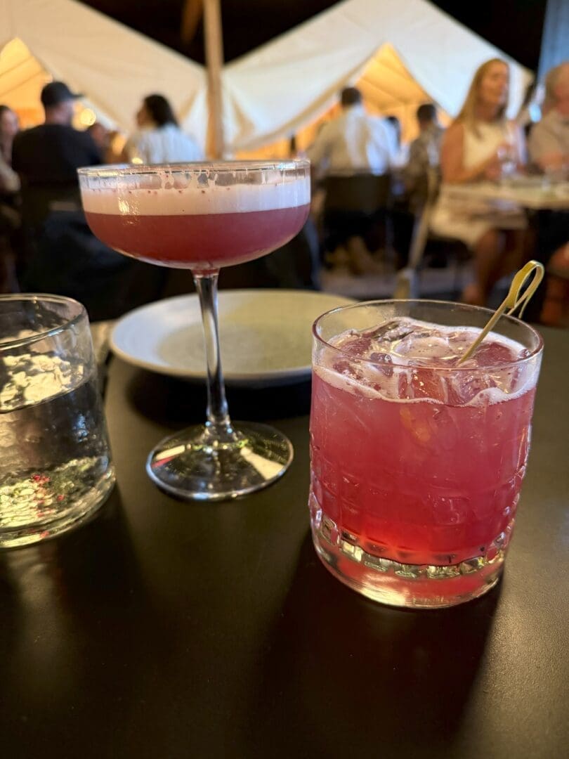 Two Pink Cocktails Sit on a Dark Table One is in a Coupe Glass with Foam on Top the Other in a Short Glass with Ice and a Cocktail Pick a Glass of Water an Empty Plate and People Dining Inside a Warmly Lit Tented Space Are Visible in the Background