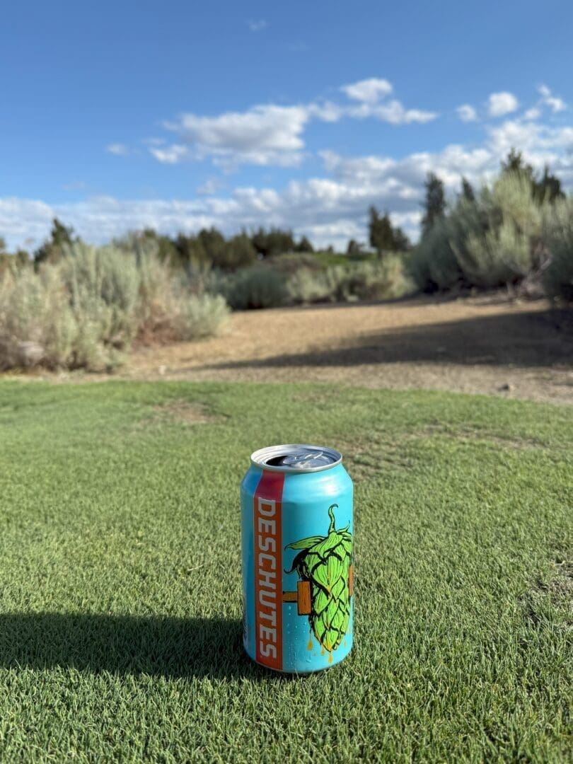 a Blue Deschutes Beer Can with a Large Green Hop Illustration and Bold Orange Lettering Stands on Green Grass at Pronghorn Golf Course in the Background a Sandy Course Weaves Among Shrubs and Trees Beneath a Partly Cloudy Blue Sky