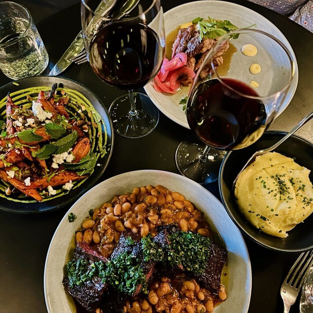 Overhead View of a Table Set with Two Glasses of Red Wine a Plate of Stewed Beans with Braised Meat and Green Sauce a Bowl of Creamy Mashed Potatoes a Dish of Roasted Carrots Topped with Mint and Feta and a Plate of Sliced Meat with Pickled Onions and Greens