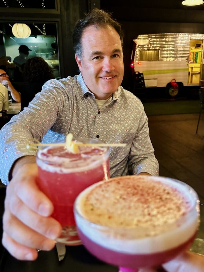 a Smiling Man in a Patterned Shirt Sits at a Table Holding Up a Pink Cocktail with a Garnish Another Hand in the Foreground Holds a Similar Pink Drink Capturing the Lively Atmosphere of Bend Restaurants with String Lights and a Shiny Food Truck Behind