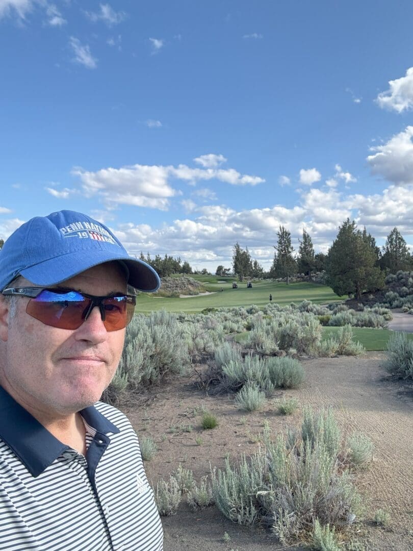 a Man in a Blue Cap Sunglasses and Striped Polo Shirt Stands in the Foreground of Pronghorn Golf Course with Green Grass Sandy Patches Low Shrubs and Distant Trees Under a Bright Blue Sky with Scattered White Clouds