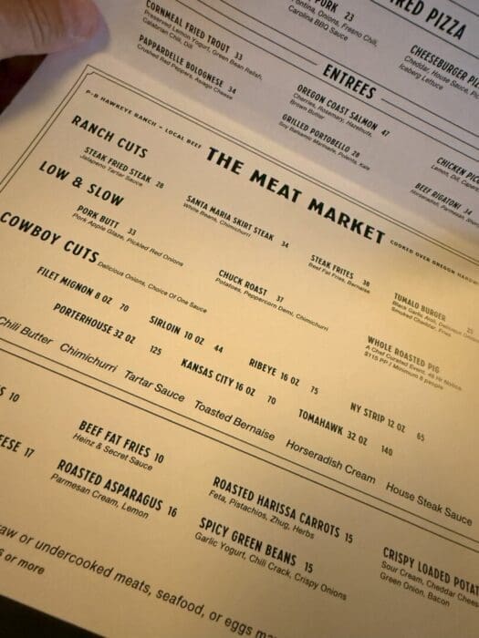 Hawkeye and huckleberry menu a Close up of a Restaurant Menu Titled the Meat Market Lists Various Steak and Meat Options Including Ranch Cuts Cowboy Cuts Low Slow and Steak Frites Each Item Has a Description and Price the Menu is Viewed at an Angle with Yellowish Lighting