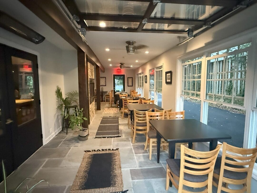 a Cozy Modern Dining Area at Market Table Bistro Features Wooden Chairs and Black Tables in a Narrow Room Large Windows Fill the Space with Natural Light Highlighting Potted Plants Wall Art Rugs on the Stone Floor and Glass Doors at Both Ends