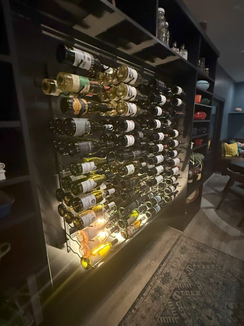 a Wall Mounted Wine Rack at Market Table Bistro Holds Dozens of Wine Bottles Arranged Horizontally on Sleek Black Metal Shelves Dim Lighting Shelves with Glassware and Bowls and a Patterned Rug Complete the Inviting Wooden Floored Room
