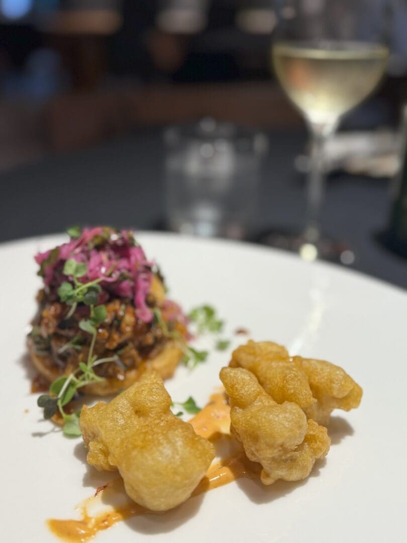 a White Plate at Market Table Bistro Holds Two Golden Crispy Fried Pieces Beside a Drizzle of Orange Sauce in the Background Shredded Meat is Topped with Purple Slaw and Microgreens While Wine and Water Glasses Sit Blurred in the Distance