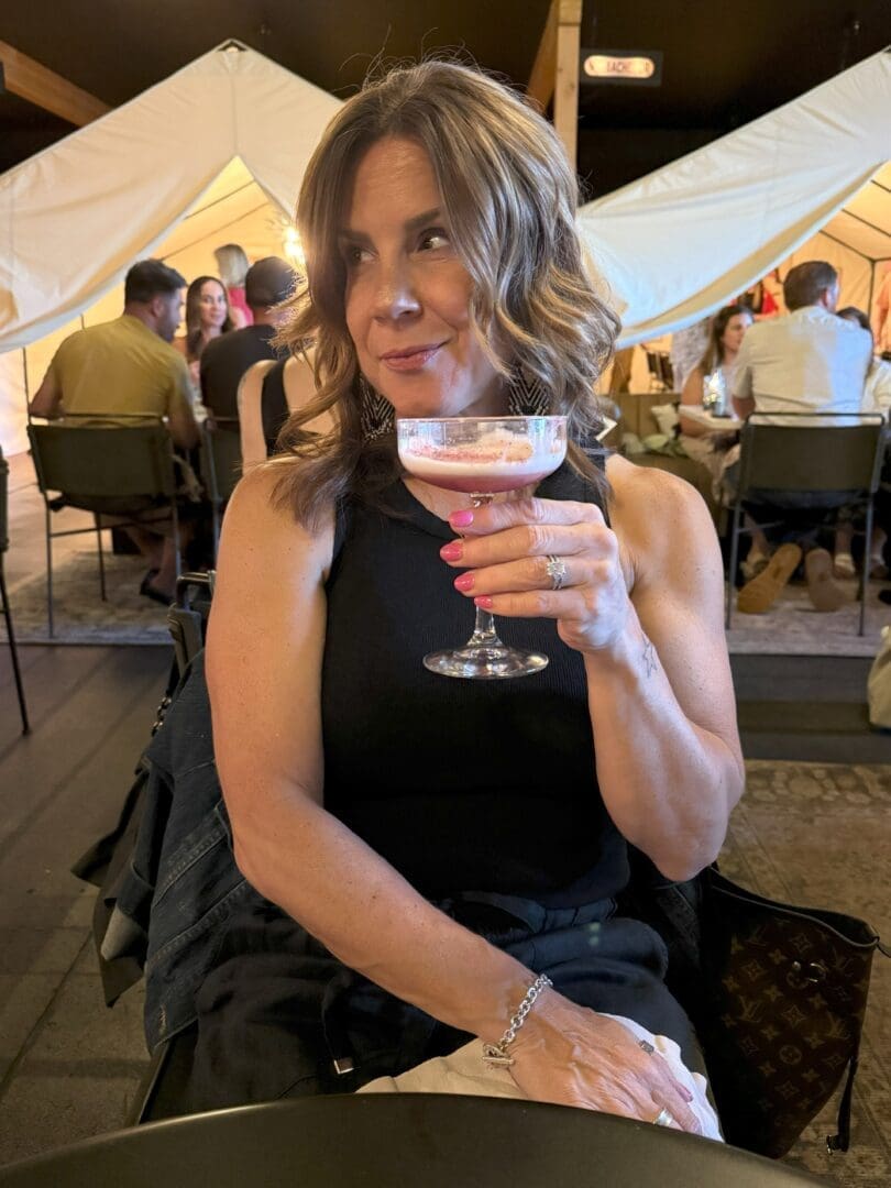 a Woman with Wavy Brown Hair in a Black Sleeveless Top Sits at a Table Holding a Pink Cocktail in a Coupe Glass Looking Thoughtfully to Her Left Behind Her People Sit at Tables in a Warmly Lit Tented Setting Suggesting a Social Gathering
