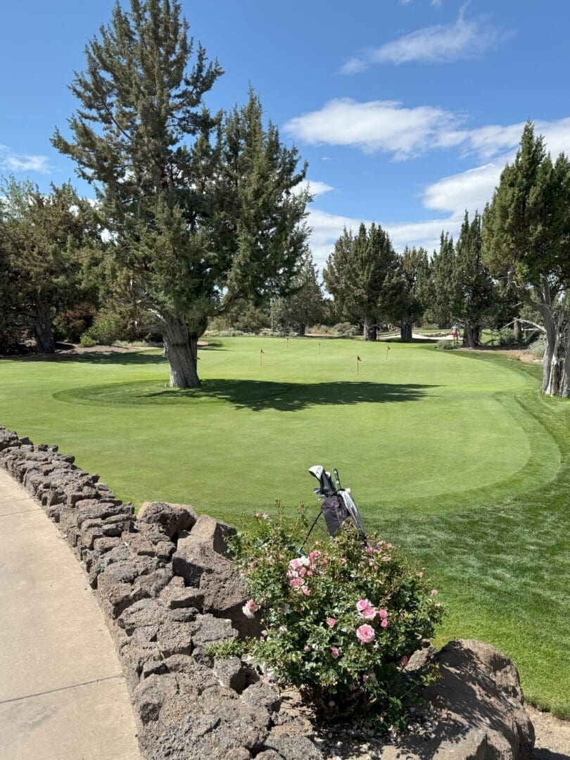 a Golf Green at Pronghorn Golf Course is Bordered by Trees and a Stone Wall with a Golf Bag Leaning Against It Pink Flowers Bloom in the Foreground While Sunlight Filters Through Partly Cloudy Skies Onto the Well kept Grass and Flags