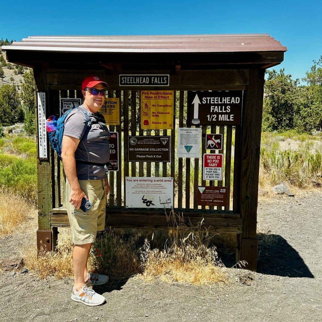 a Person in a Red Cap Sunglasses Blue Shirt Beige Shorts and Sneakers Stands Smiling Beside a Steelhead Falls Trail Signboard Filled with Notices and Maps They Carry a Backpack Amid Sunny Surroundings of Dry Grass and Sparse Trees