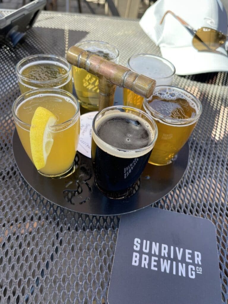 Sunriver Brewing Beer Flight a Round Metal Tray on an Outdoor Metal Table Holds Six Small Glasses of Beer One with a Lemon Slice the Tray Has a Brass Handle a Sunriver Brewing Co Coaster and Sunglasses Hint at the Great Breweries in Bend Oregon