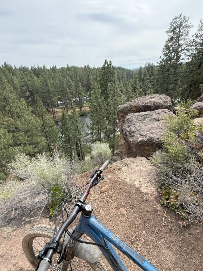 a Mountain Bike Rests on Rimrock Trail at the Edge of a Rocky Cliff Overlooking a Forest of Tall Pine Trees and a Small River Below the Sky is Overcast with Bushes and Shrubs Lining the Trail in the Foreground