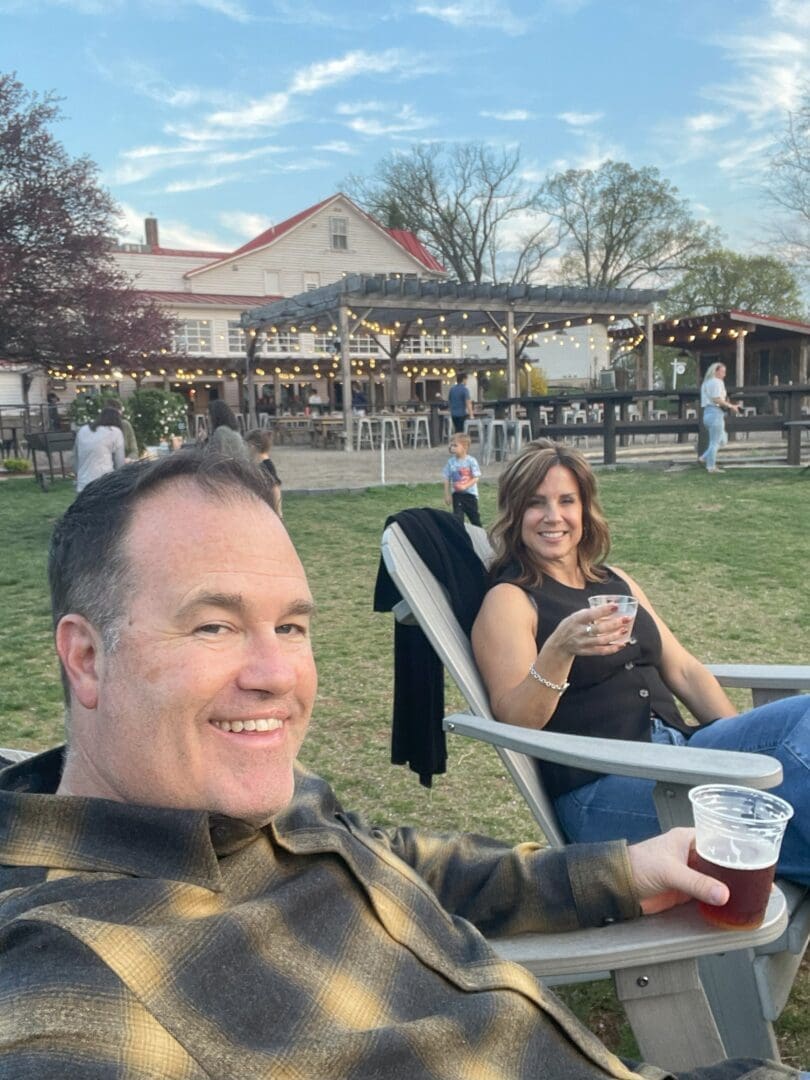 a Man and Woman Sit on Lawn Chairs Outdoors Smiling and Holding Drinks Behind Them is a Large Farmhouse Style Building with String Lights People Walking and Treesall Creating a Relaxed Vibe That Invites You to Work with Us in This Cheerful Setting