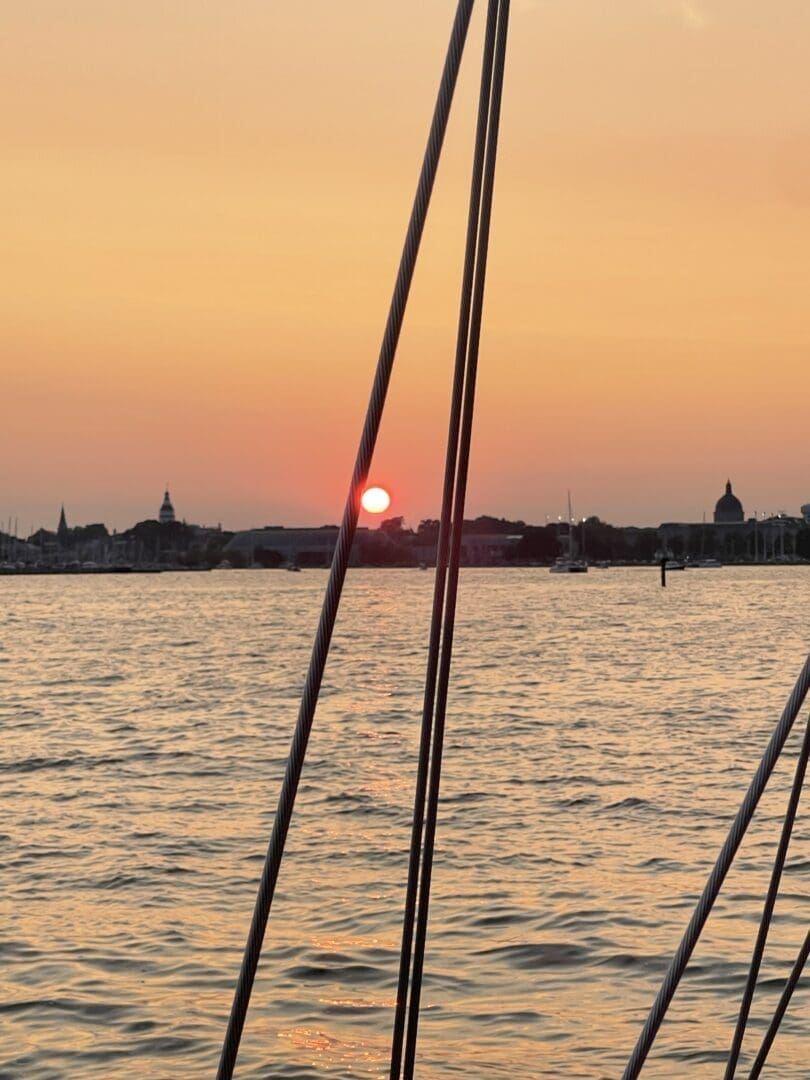 a Vivid Sunset over Calm Water in Annapolis Maryland with the Orange Sun Low in the Sky Sailboat Rigging Lines Cross the Foreground While Silhouetted Buildings and Boats Line the Distant Shore Under a Soft Orange and Yellow Sky