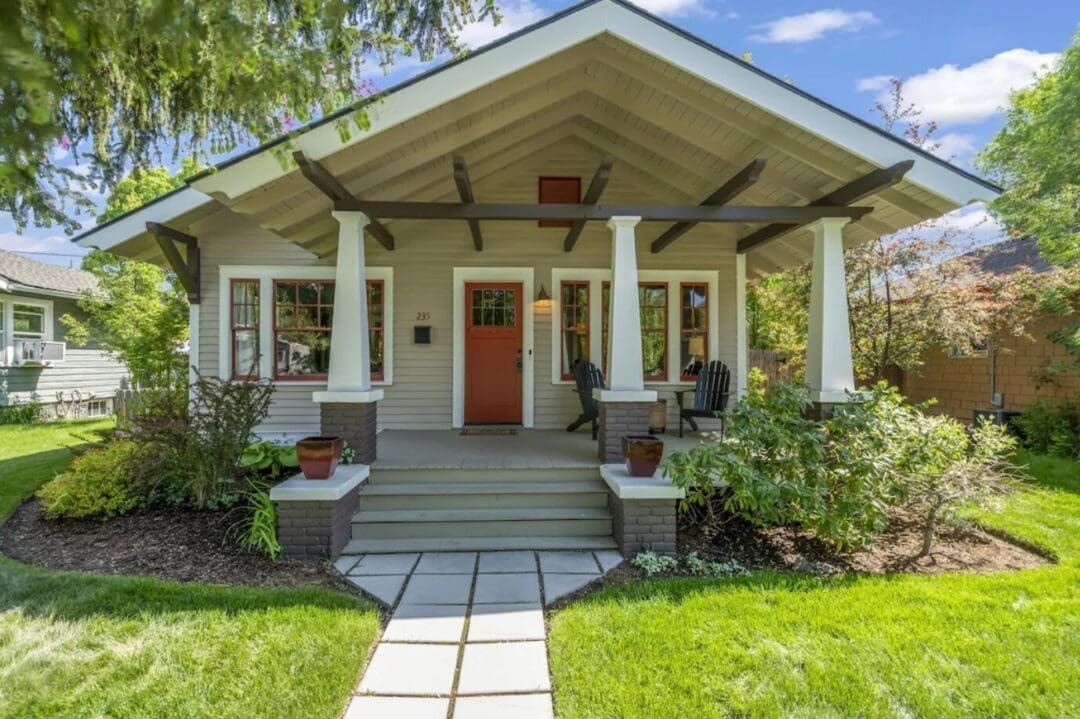 A charming craftsman-style house—an ideal option if you’re wondering where to stay in Bend, Oregon. Enjoy the front porch with white columns, a red door, brown trim, and lush greenery all around under a blue sky dotted with white clouds.
