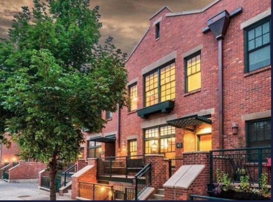 a Row of Modern Brick Townhouses at Dusk with Large Lit Windows and Black Railings Lining Small Front Stepsthese Inviting Homes Evoke the Charm Youd Seek when Deciding Where to Stay in Bend Oregon a Leafy Tree Completes the Picturesque Scene