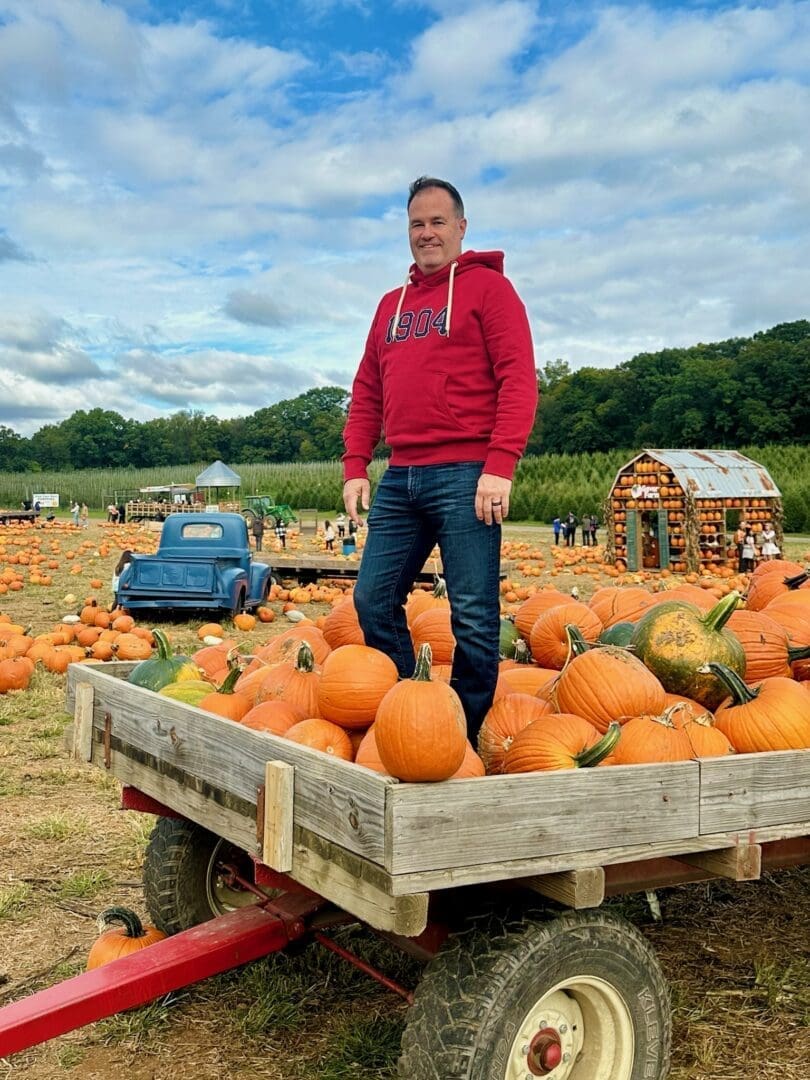 a Man in a Red Hoodie Stands Smiling on a Wooden Wagon Filled with Pumpkins at Gaver Farms Large Pumpkin Patch Scattered Pumpkins a Blue Vintage Truck and a Barn Like Structure Appear in the Background Under a Partly Cloudy Sky
