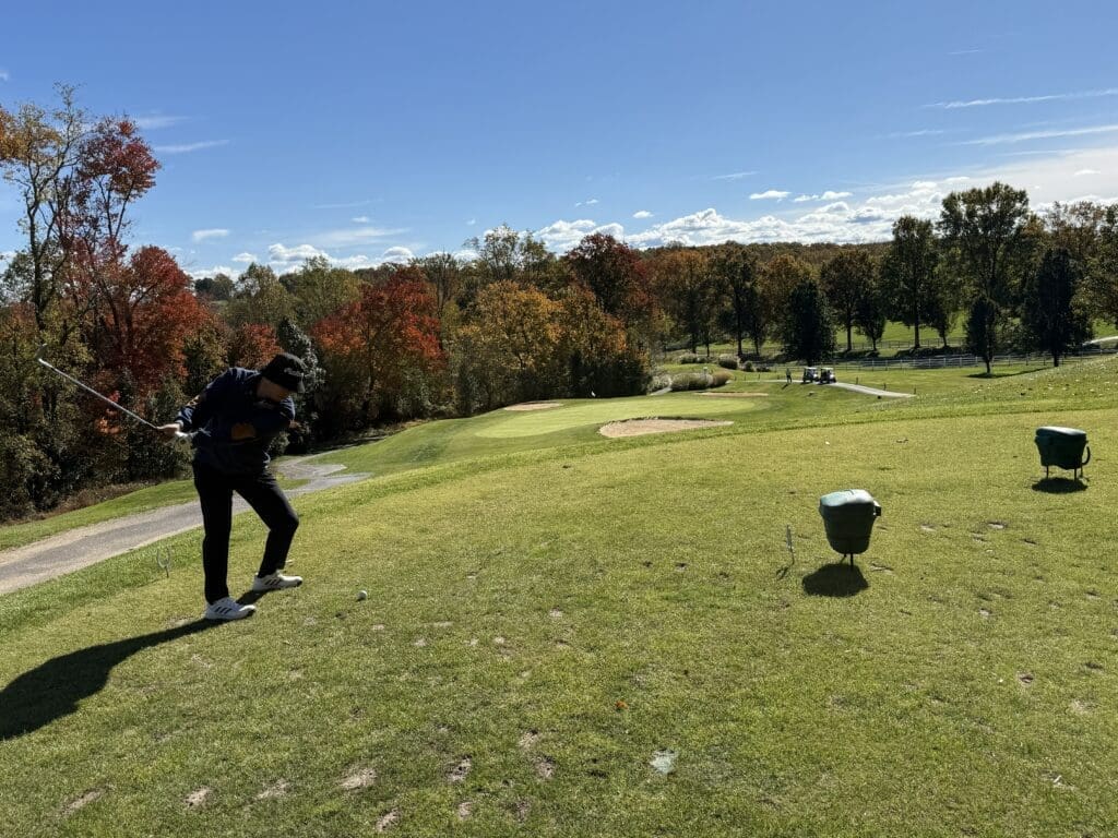 a Golfer is Mid Swing on a Tee Box at the Links at Challedon Surrounded by Green Grass and Autumn Trees with Red Yellow and Orange Leaves Under a Clear Blue Sky Two Green Baskets Are Nearby with Distant Golf Carts in the Background