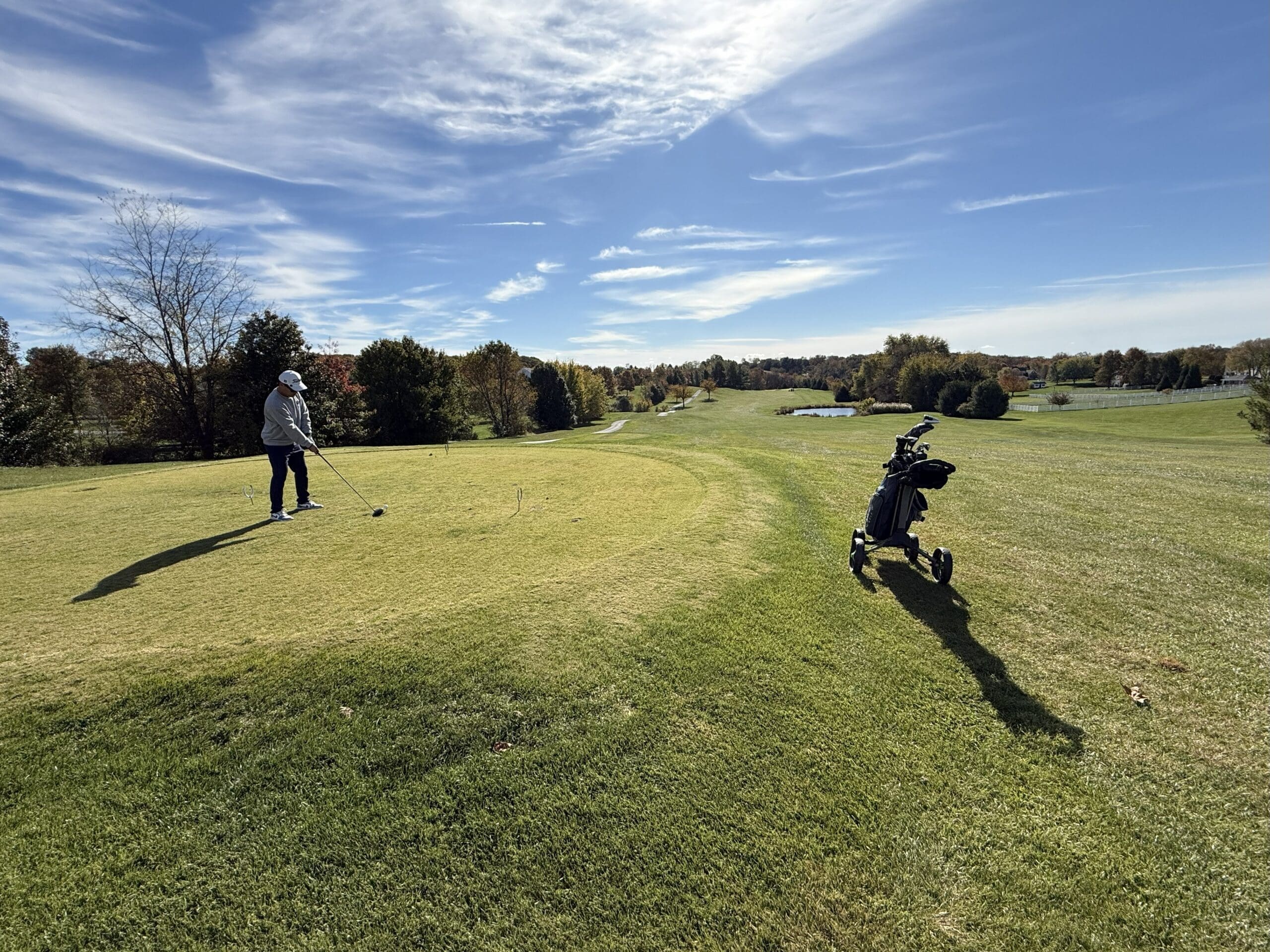 a Golfer Stands on the Tee at the Links at Challedon Preparing to Swing on a Sunlit Course with Autumn Trees in the Distance a Pushcart with Clubs Casts a Long Shadow Across the Grass Under a Partly Cloudy Blue Sky
