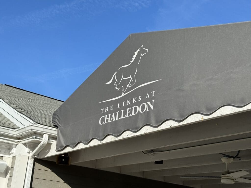 a Gray Awning Displays a White Outline of a Running Horse Above the Words the Links at Challedon the Awning Covers Part of a Building with White Trim Highlighting the Distinctive Entrance to the Links at Challedon Under a Clear Blue Sky