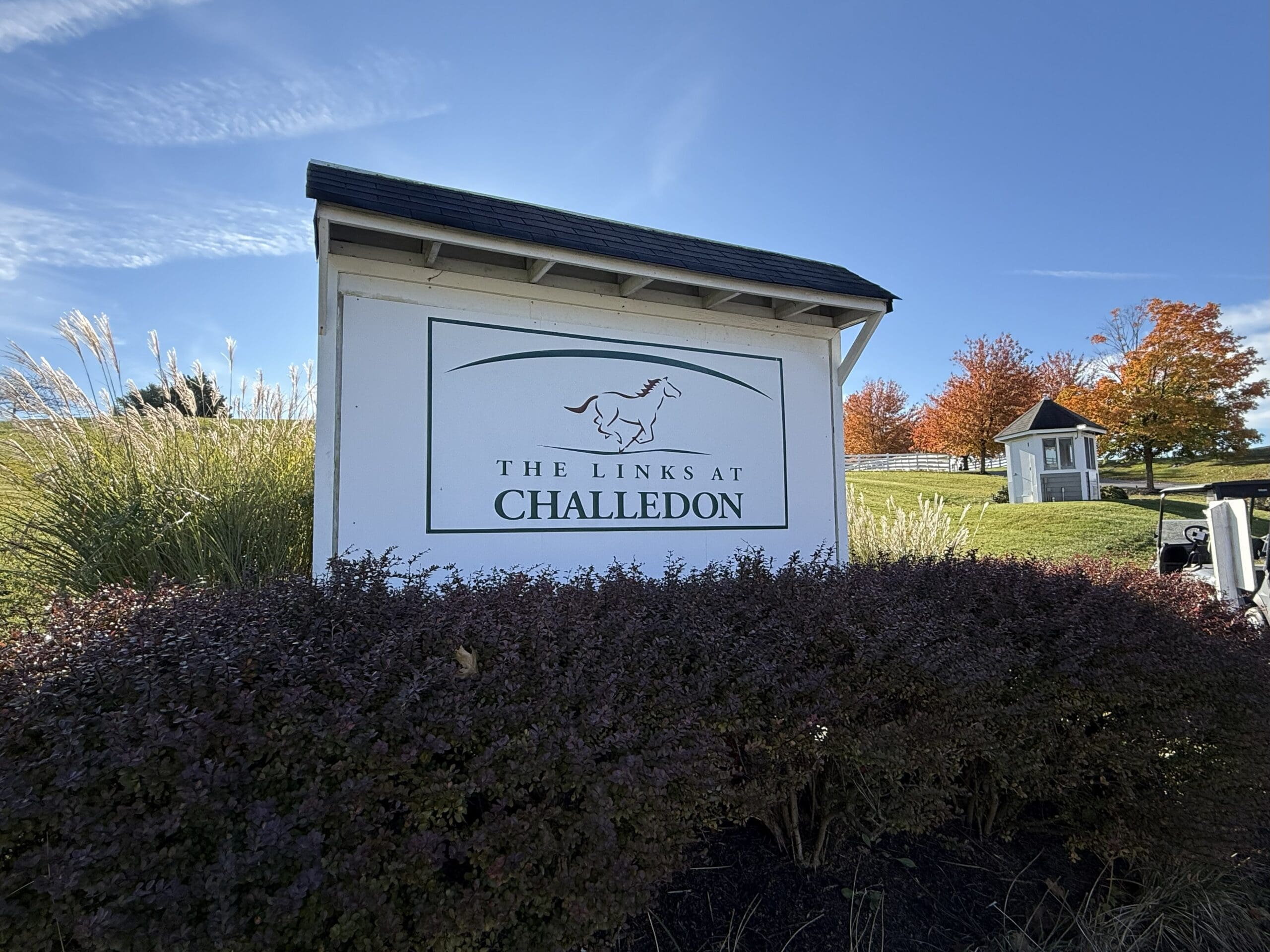 a White Sign with a Horse Logo and Text Reading the Links at Challedon Stands Among Shrubs and Tall Grass in Mt Airy Md in the Background Autumn Trees with Orange Leaves and a Small Shed Sit Under a Bright Blue Sky with Wispy Clouds